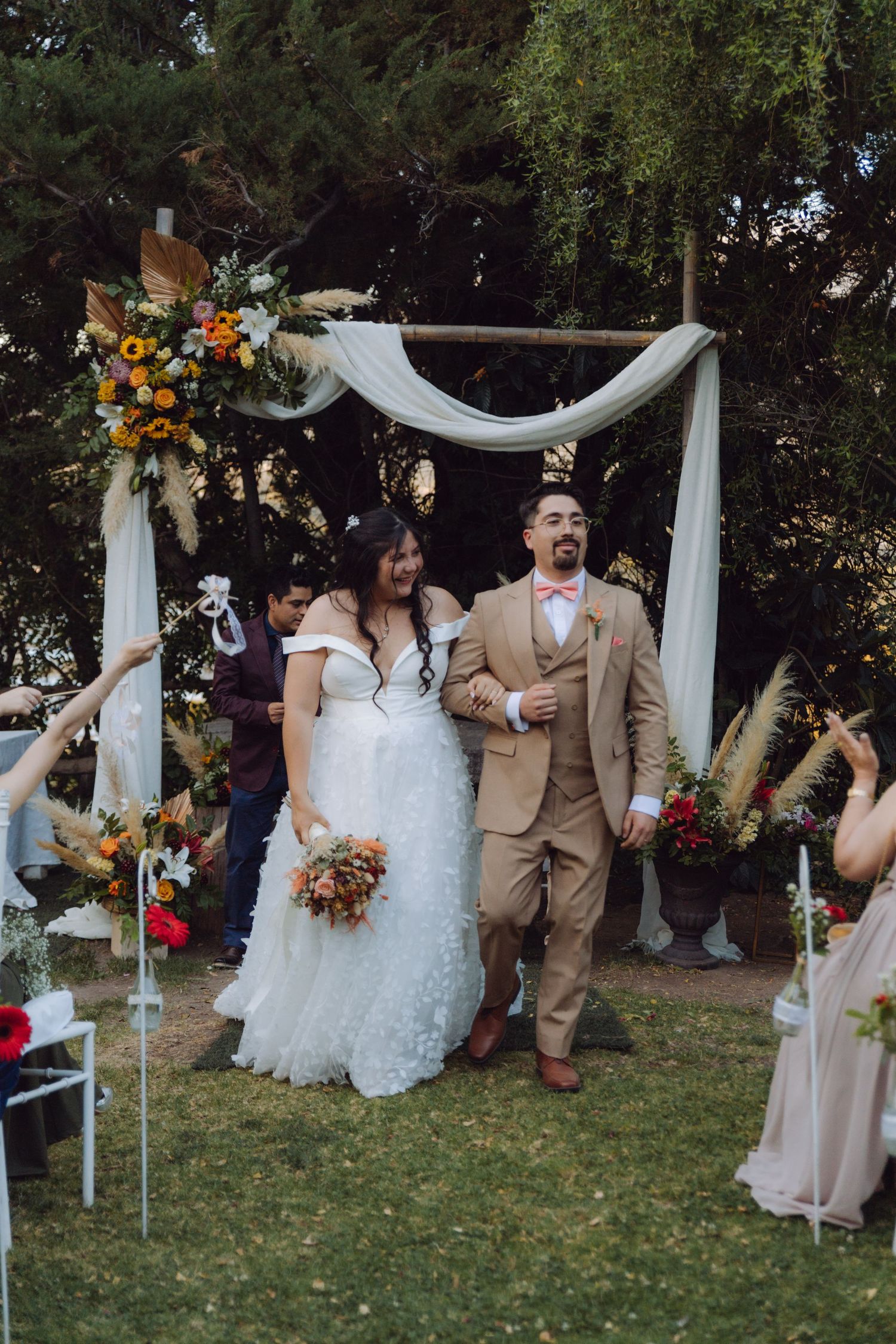 Happy bride and groom exit outdoor wedding ceremony arm in arm under rustic floral arch.