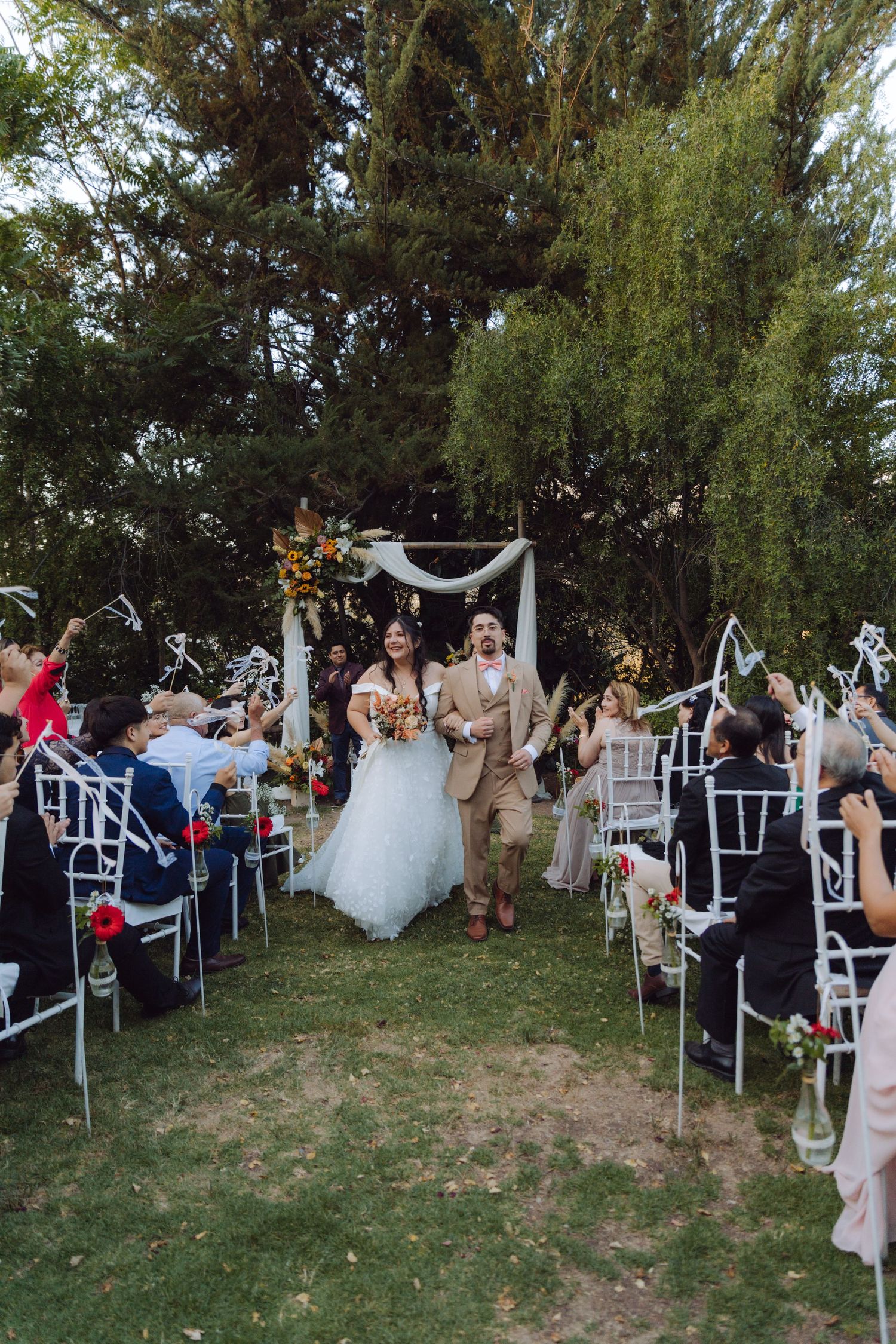 Bride and groom walk down aisle as guests wave streamers at outdoor garden wedding ceremony.