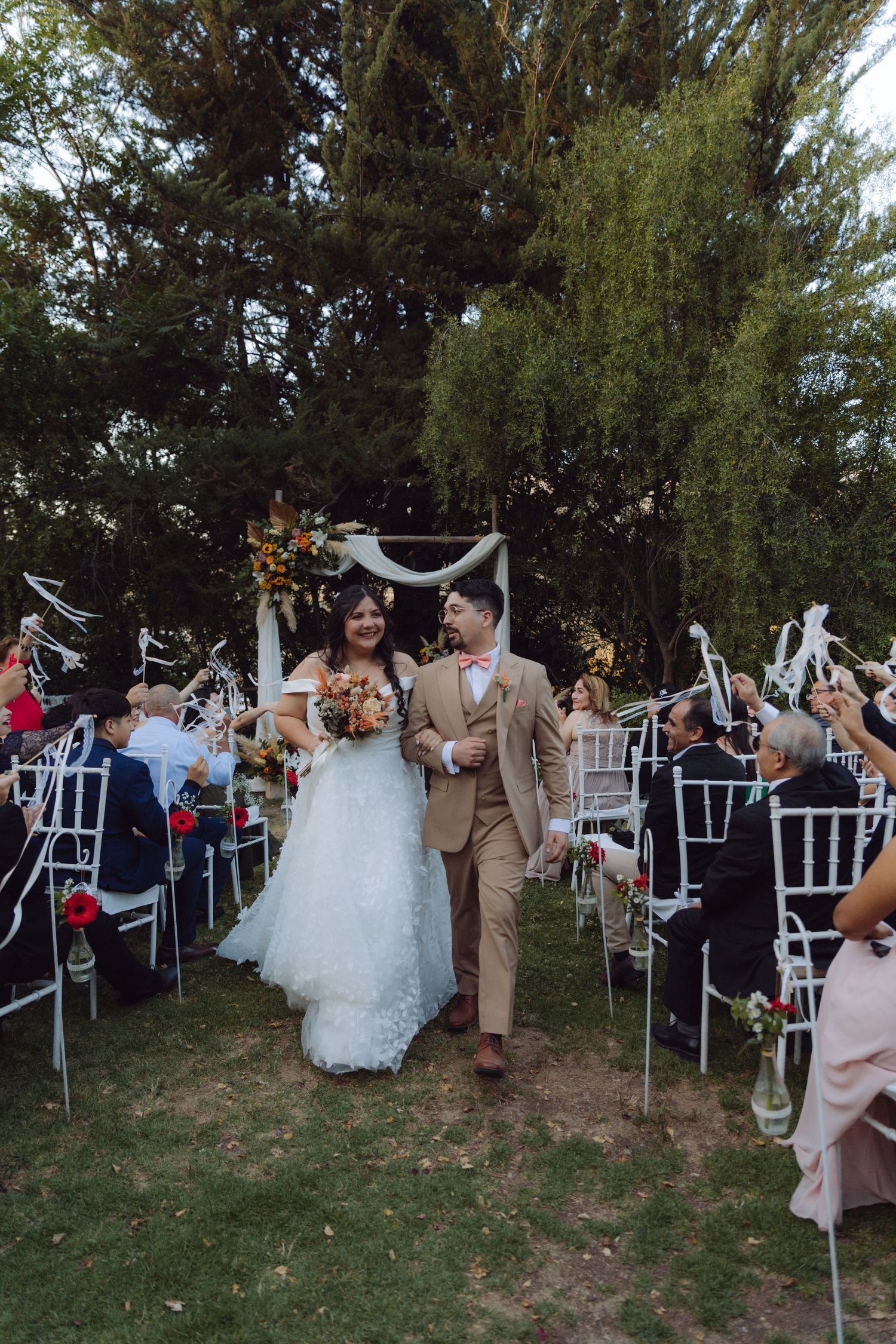 Smiling newlyweds walk down aisle as guests celebrate at outdoor garden wedding ceremony.