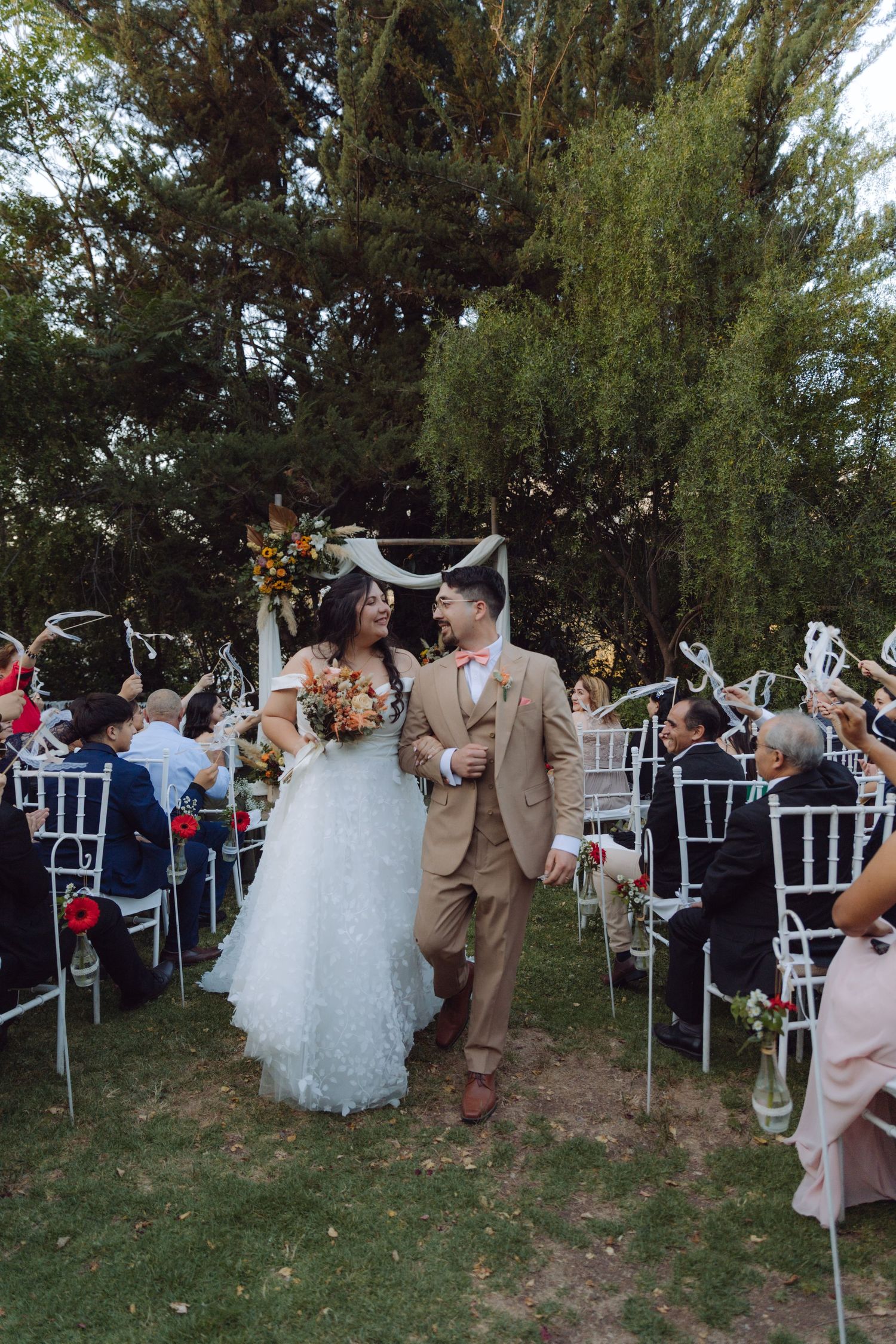 Happy newlyweds walk down outdoor aisle as guests celebrate with streamers at garden wedding ceremony.