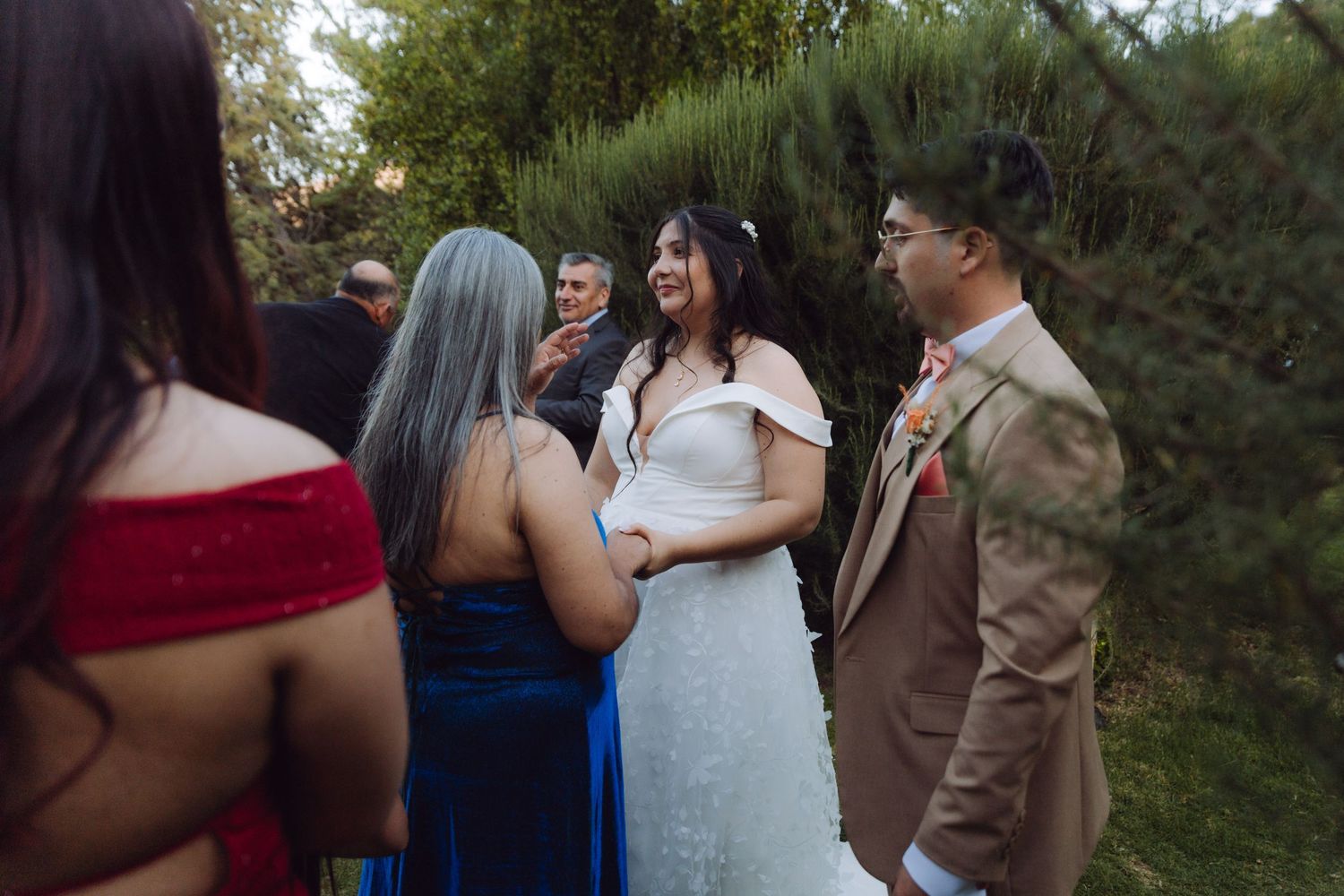 Smiling bride in white off-shoulder gown greets guests after outdoor garden wedding ceremony.