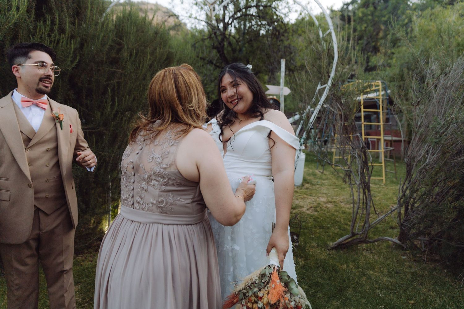 Bride in white gown laughs with guest in gray dress while groom watches at outdoor wedding.