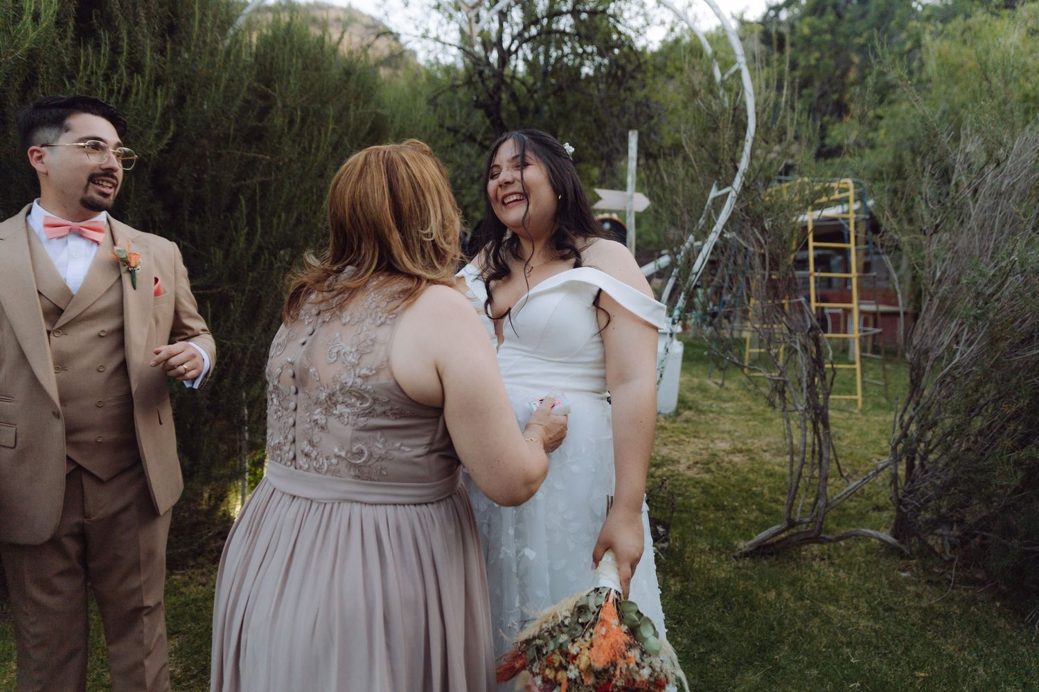 Bride joyfully embraces a guest in a gray lace dress while groom smiles at garden wedding.