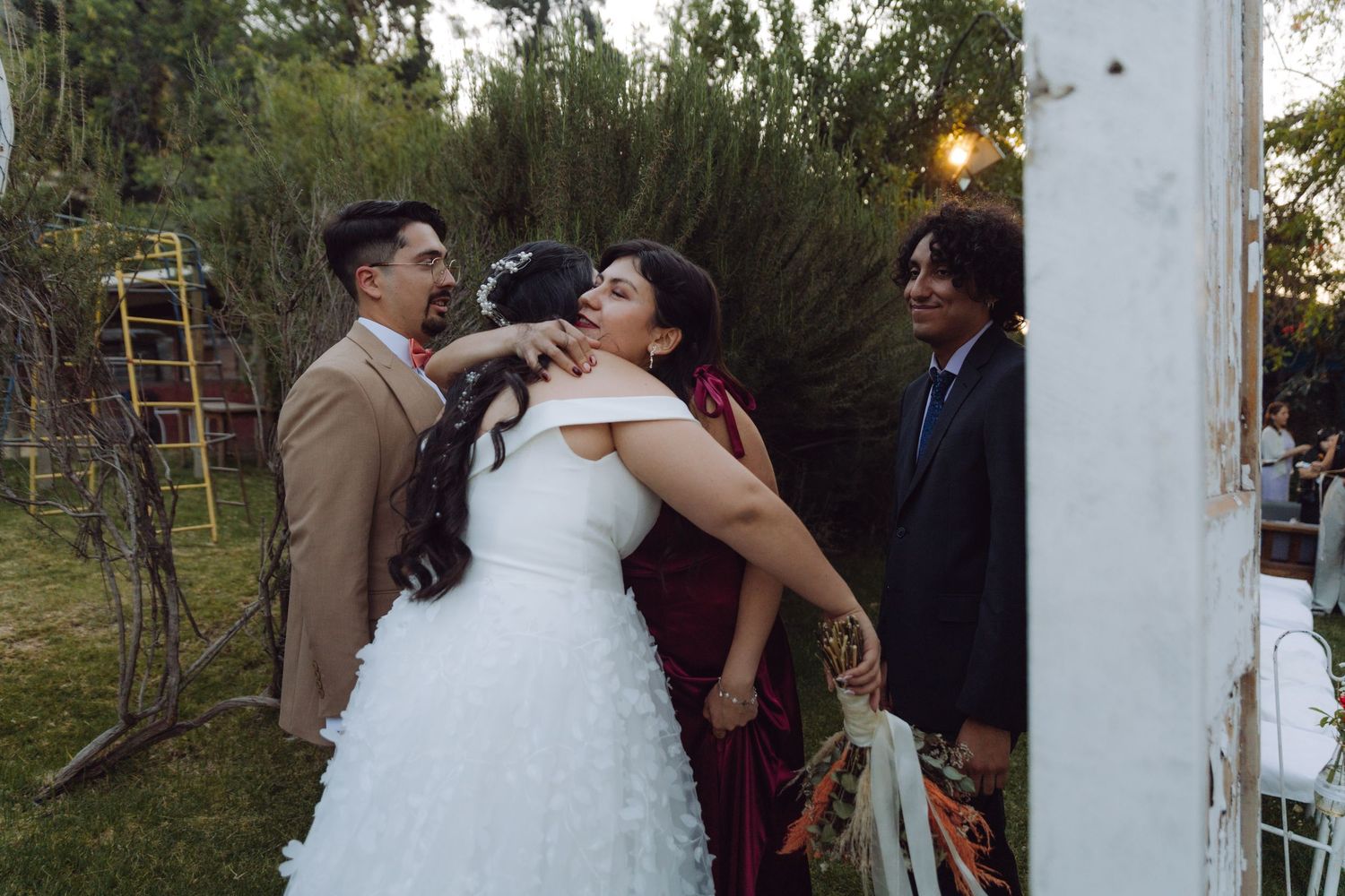 Bride in white gown receives a kiss on the cheek from a guest in red at outdoor wedding.