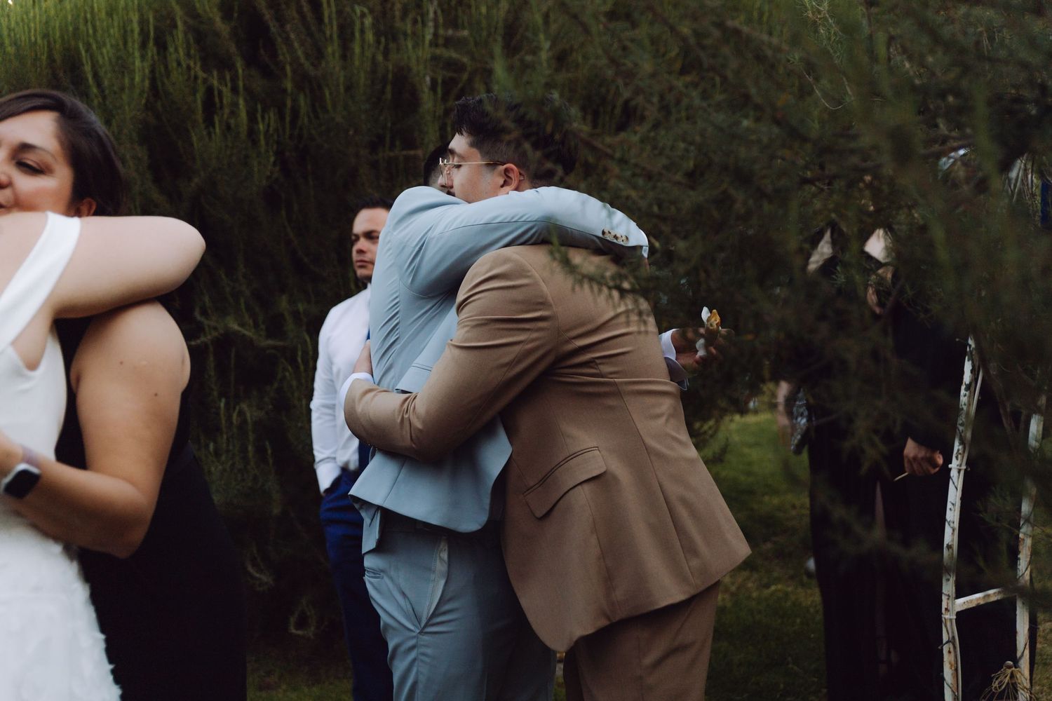 Groom in tan suit shares a warm embrace with a guest in blue at outdoor wedding reception.