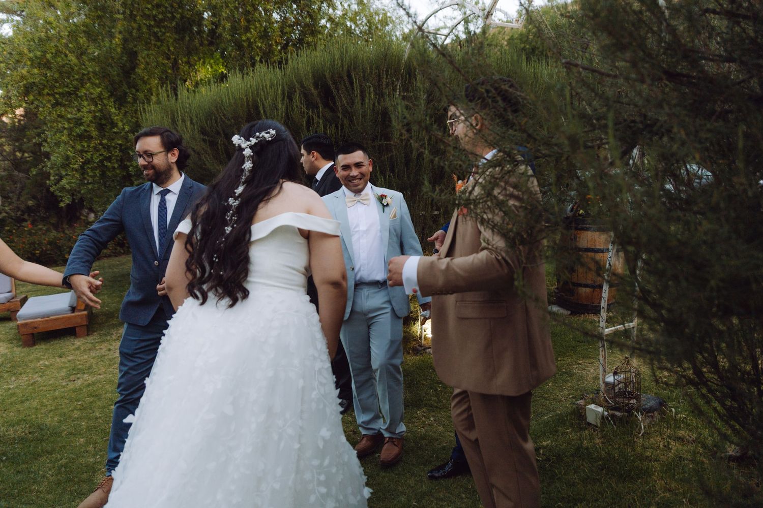 Bride and groom in tan suit greet smiling guests in a lush garden after wedding ceremony.