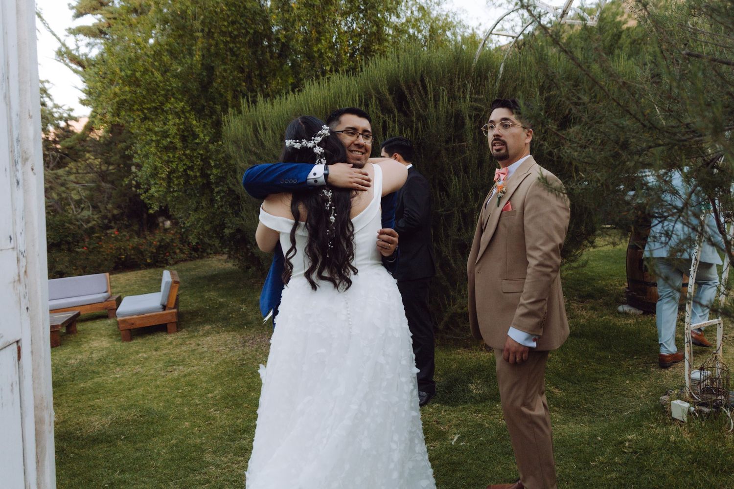 Bride and groom embrace a guest while another man stands nearby in a green garden setting.