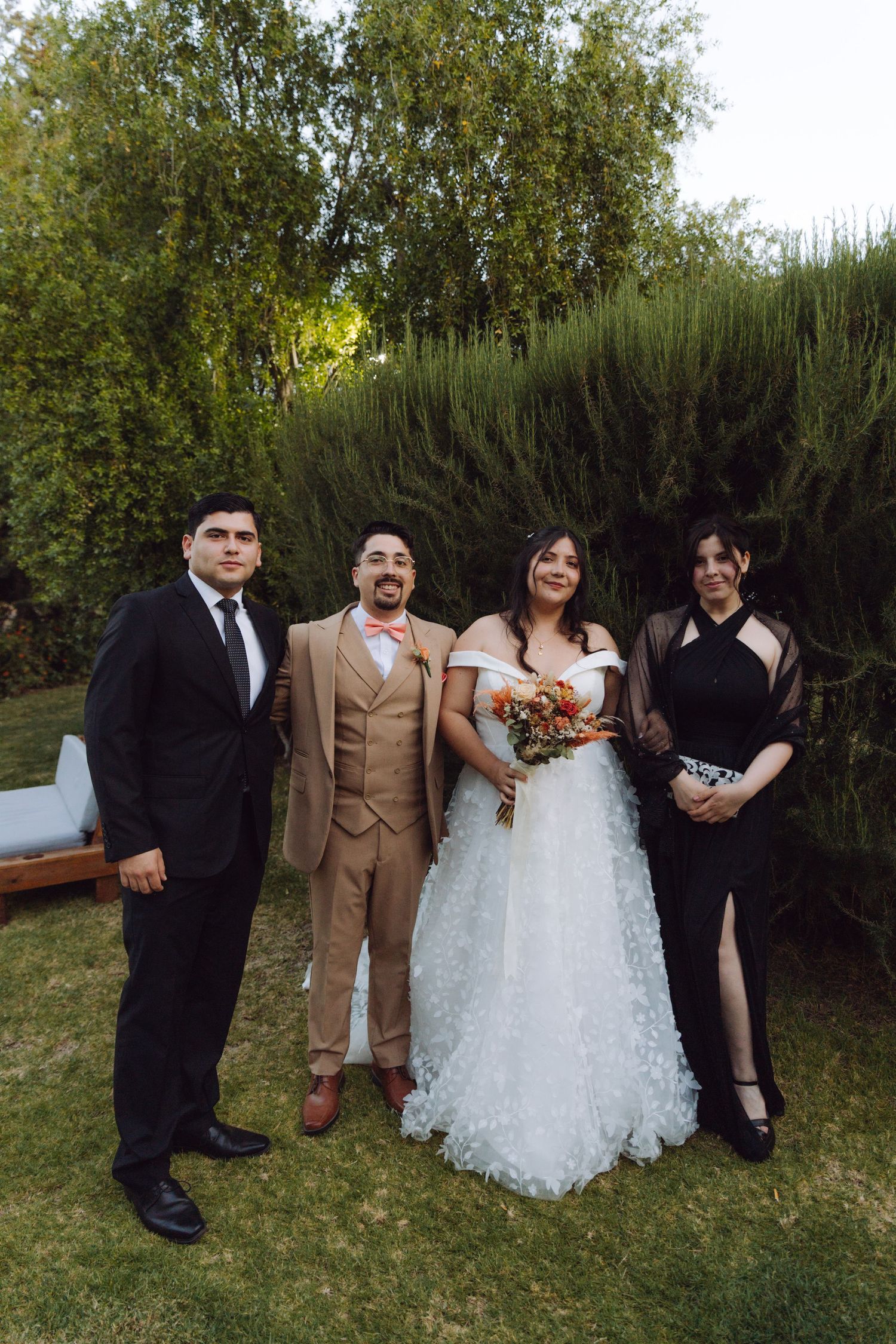 Bride and groom pose with two guests in formal attire in a lush green garden after wedding.