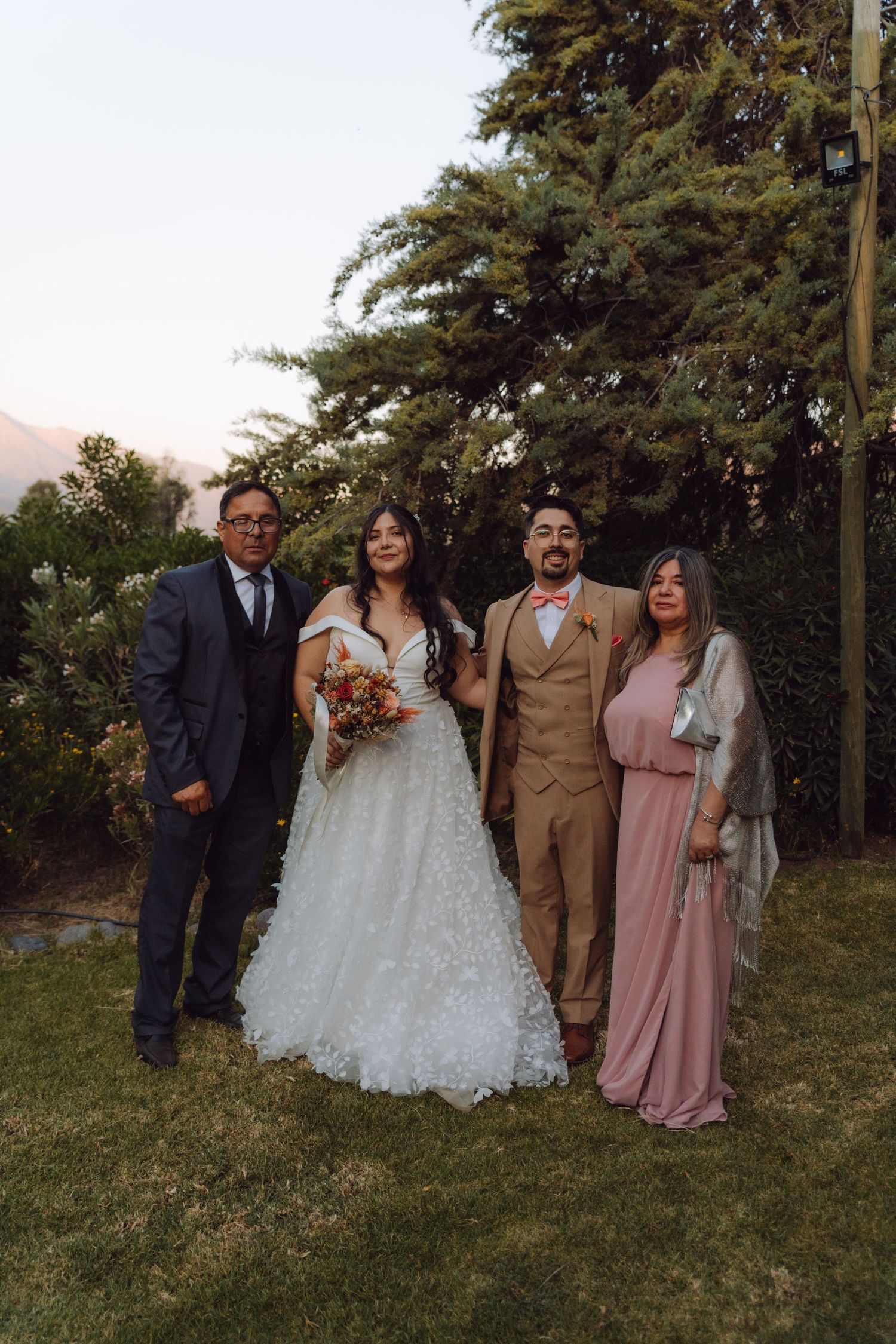 Wedding couple poses with family on green lawn, bride in white gown holding bouquet, groom in tan suit, outdoor garden setting.