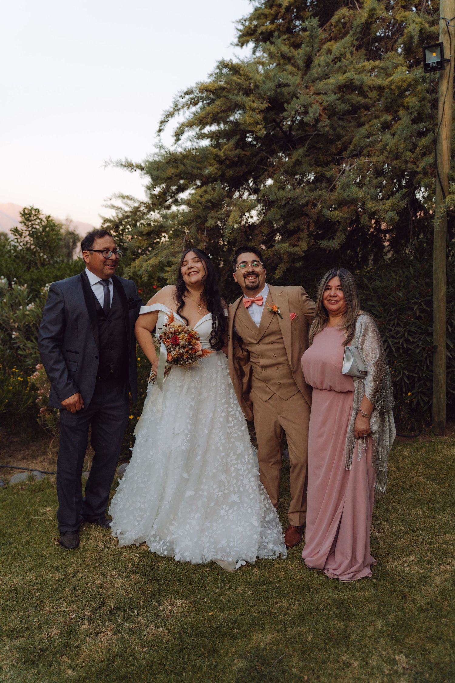 Smiling wedding party of four poses outdoors, bride in white lace gown, groom in tan suit with pink bow tie, garden backdrop.