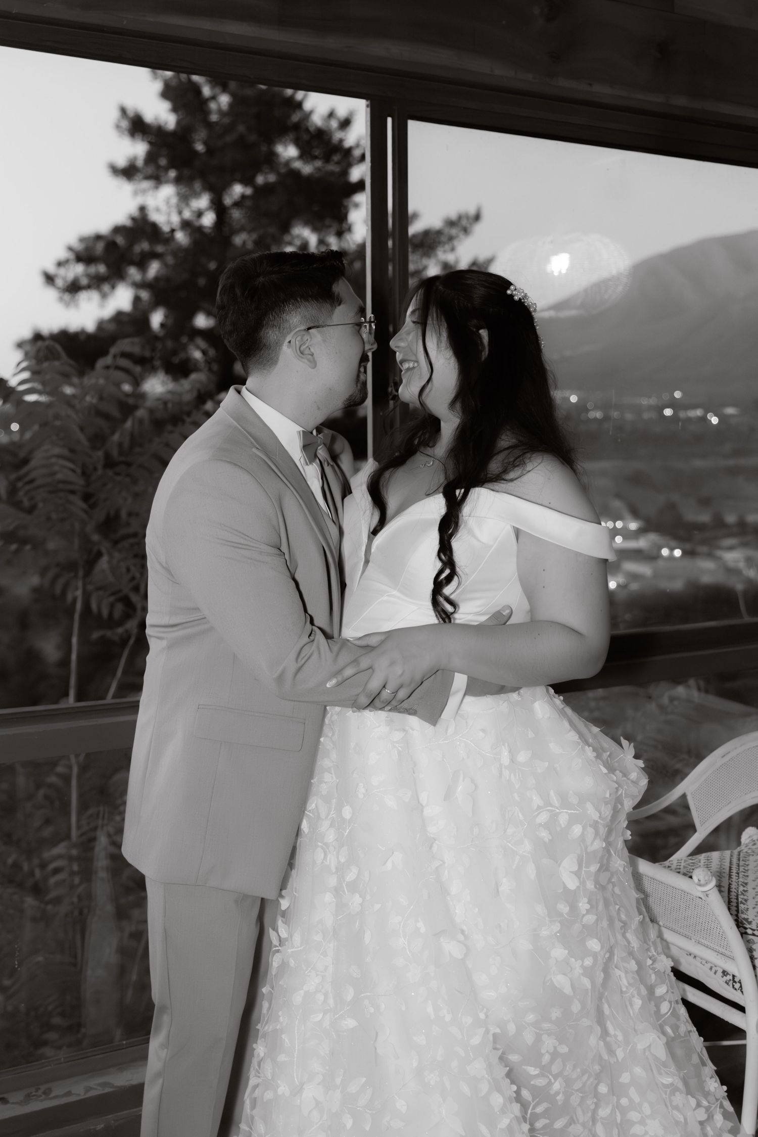 Black and white romantic portrait of bride and groom embracing near window with mountain view at twilight.