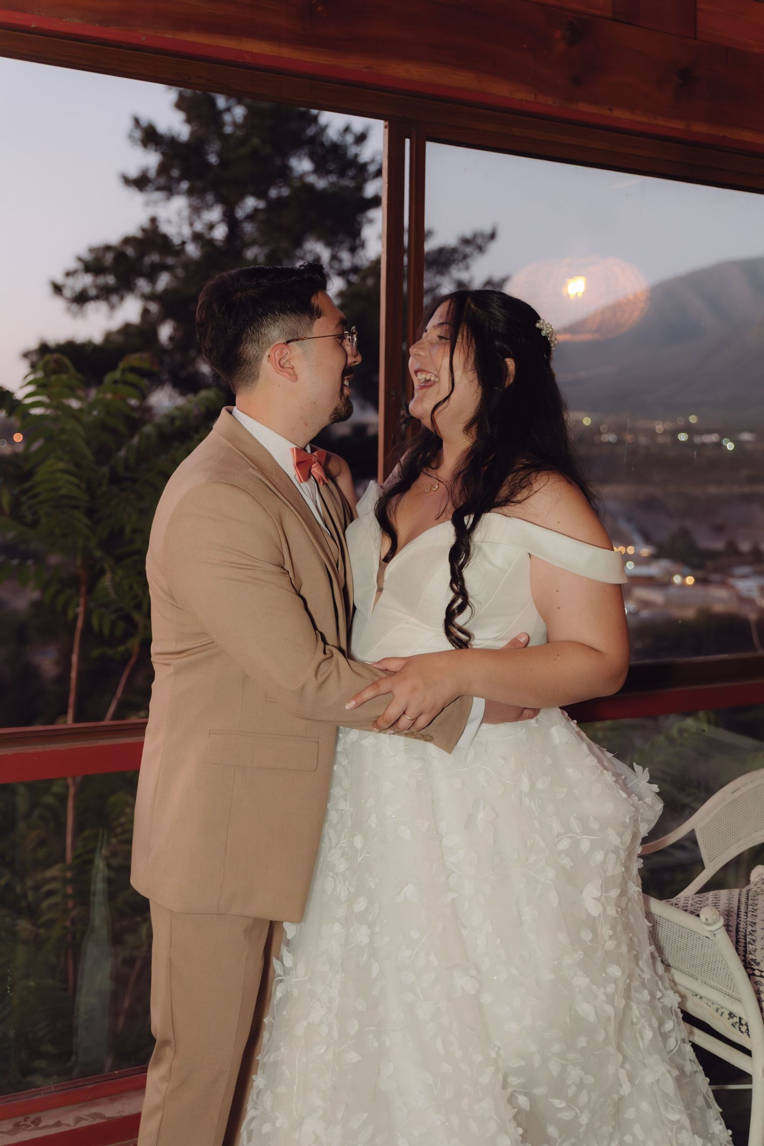 Bride and groom share a joyful moment holding hands on a balcony at sunset with mountain views.