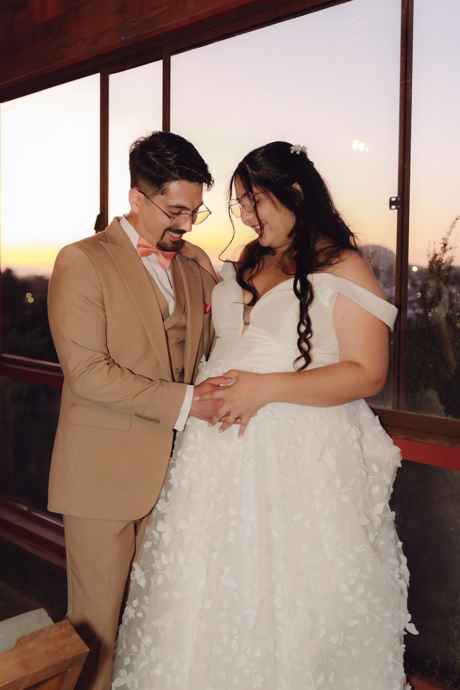 Bride and groom share a tender moment at sunset, she in a floral white gown, he in a tan suit with pink bow tie.