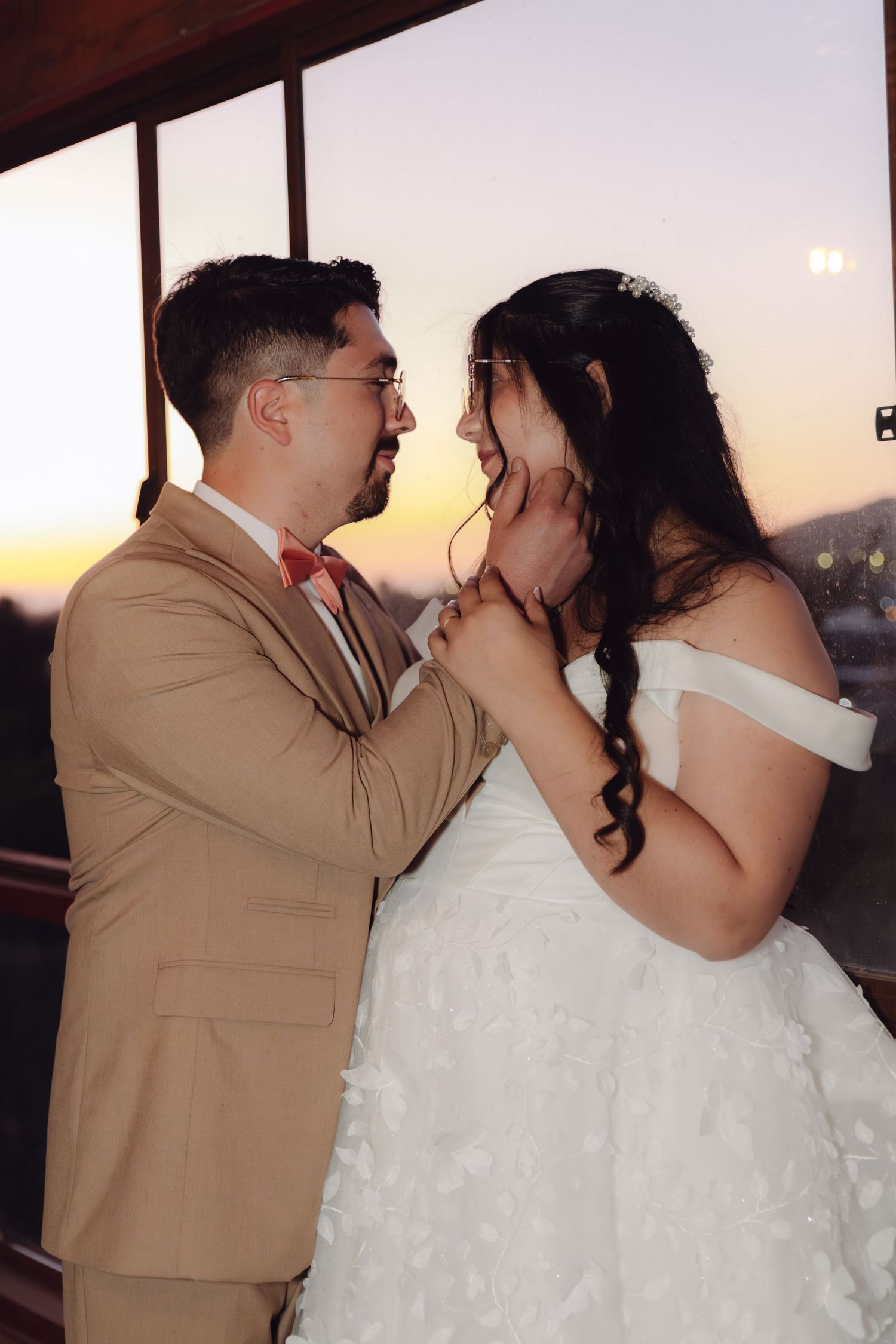 Groom tenderly cups bride's face at sunset, both smiling in romantic wedding portrait on venue balcony.