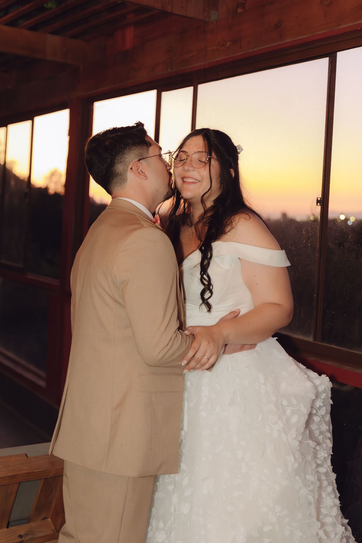 Groom kisses bride's cheek at sunset as she smiles joyfully, both embracing in wedding attire on balcony.