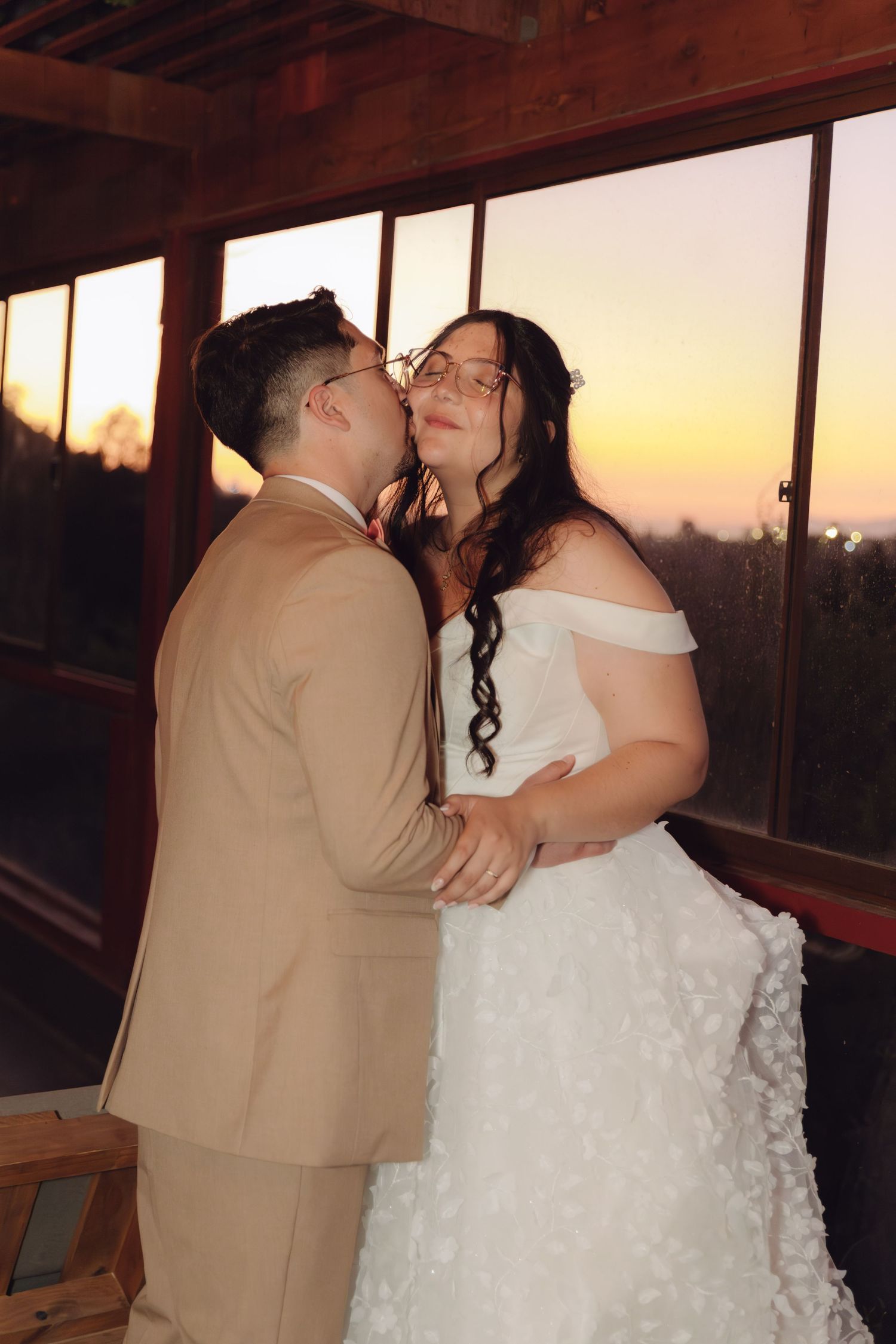 Groom kisses smiling bride on cheek during golden sunset, both holding hands in elegant wedding attire.