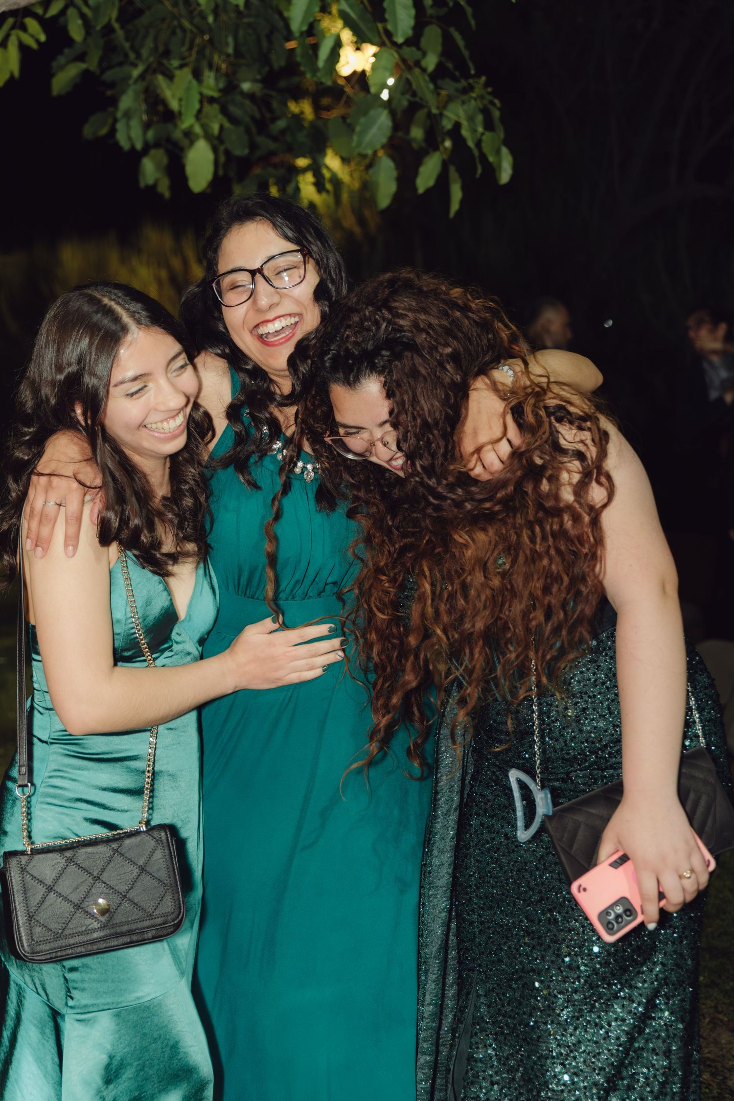 Three joyful women in emerald green dresses laughing and hugging at an outdoor wedding reception at night.
