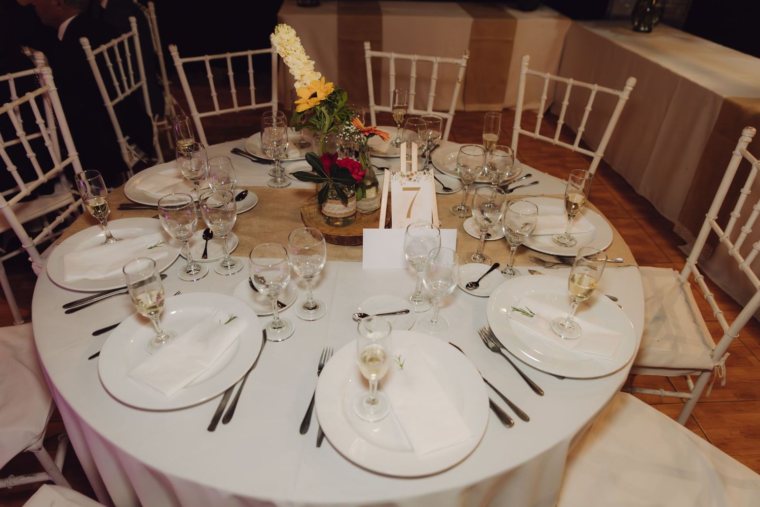 Elegant round wedding reception table set with white linens, crystal glasses, white chairs, and floral centerpiece, table number 7.