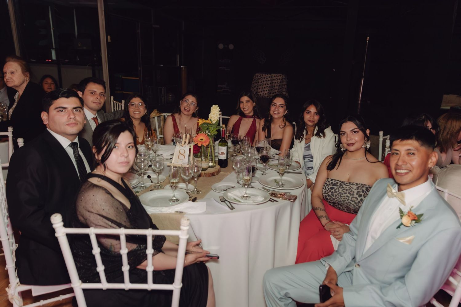 Wedding guests smile and pose together at a decorated reception dinner table with floral centerpieces and wine bottles.