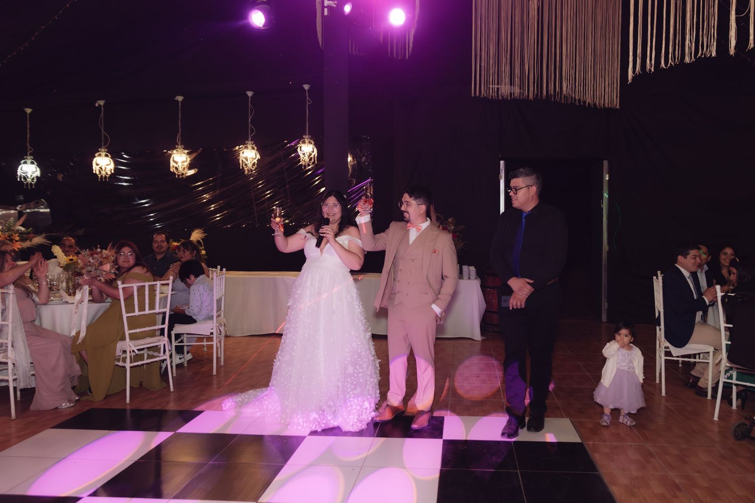 Bride and groom stand on checkered dance floor with guest during wedding reception in softly lit venue.