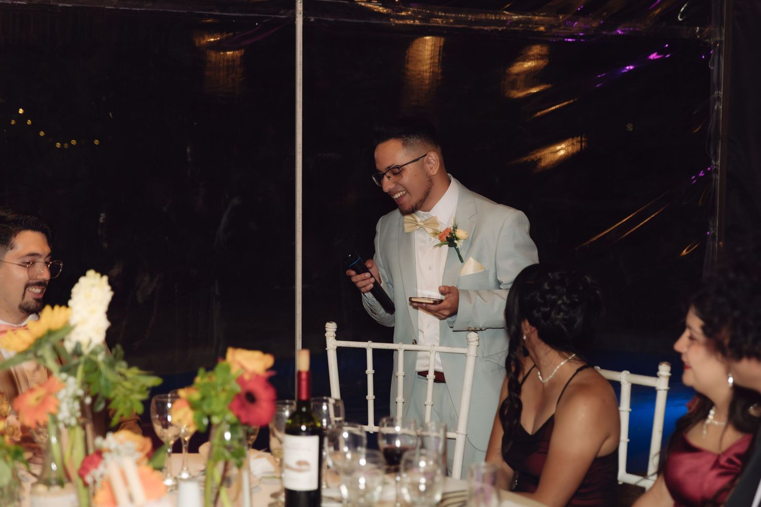 Man in light suit gives a toast speech at a wedding reception table decorated with colorful floral arrangements.