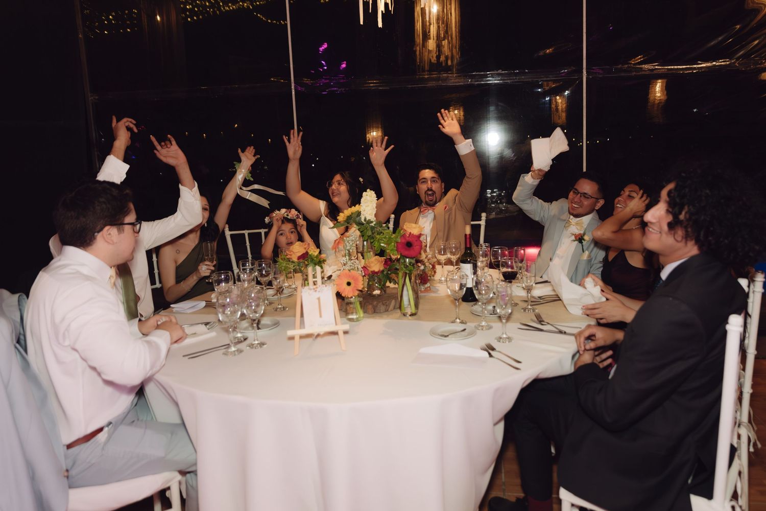 Wedding guests raising hands in celebration at a decorated reception dinner table at night.