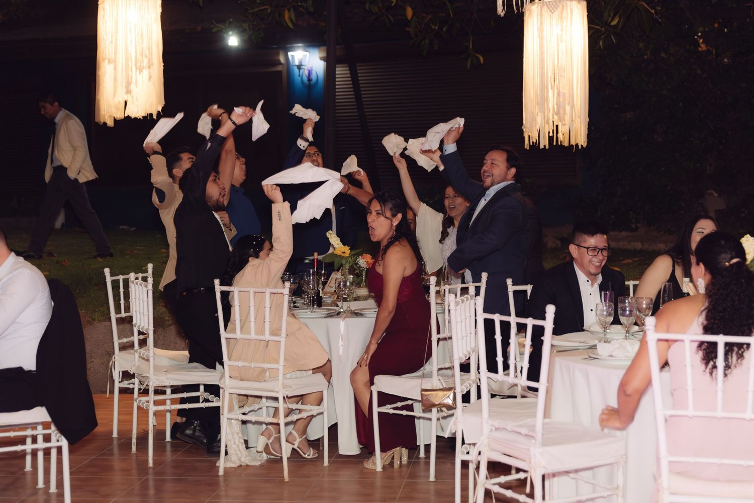 Wedding guests waving white napkins in celebration at an outdoor evening reception venue.