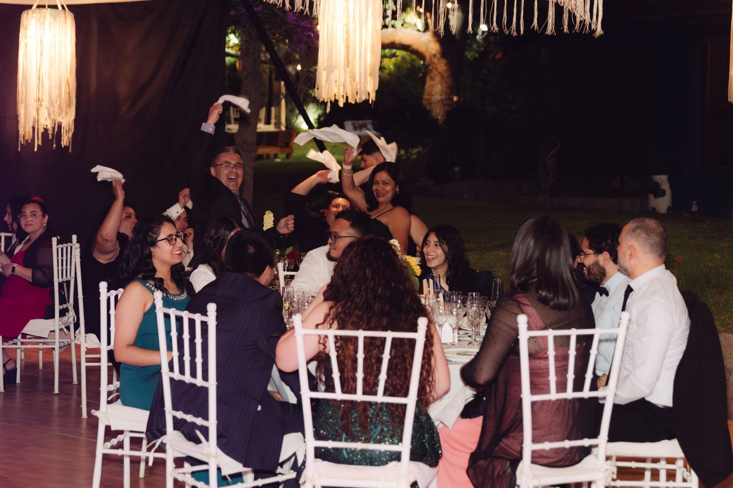 Joyful wedding guests waving napkins at a festive outdoor nighttime reception with white chairs.