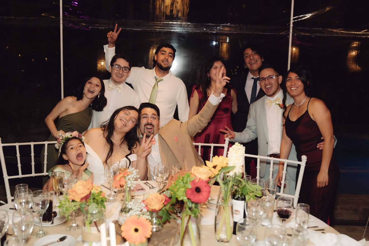 Happy wedding party group photo at decorated reception table with colorful floral centerpieces.