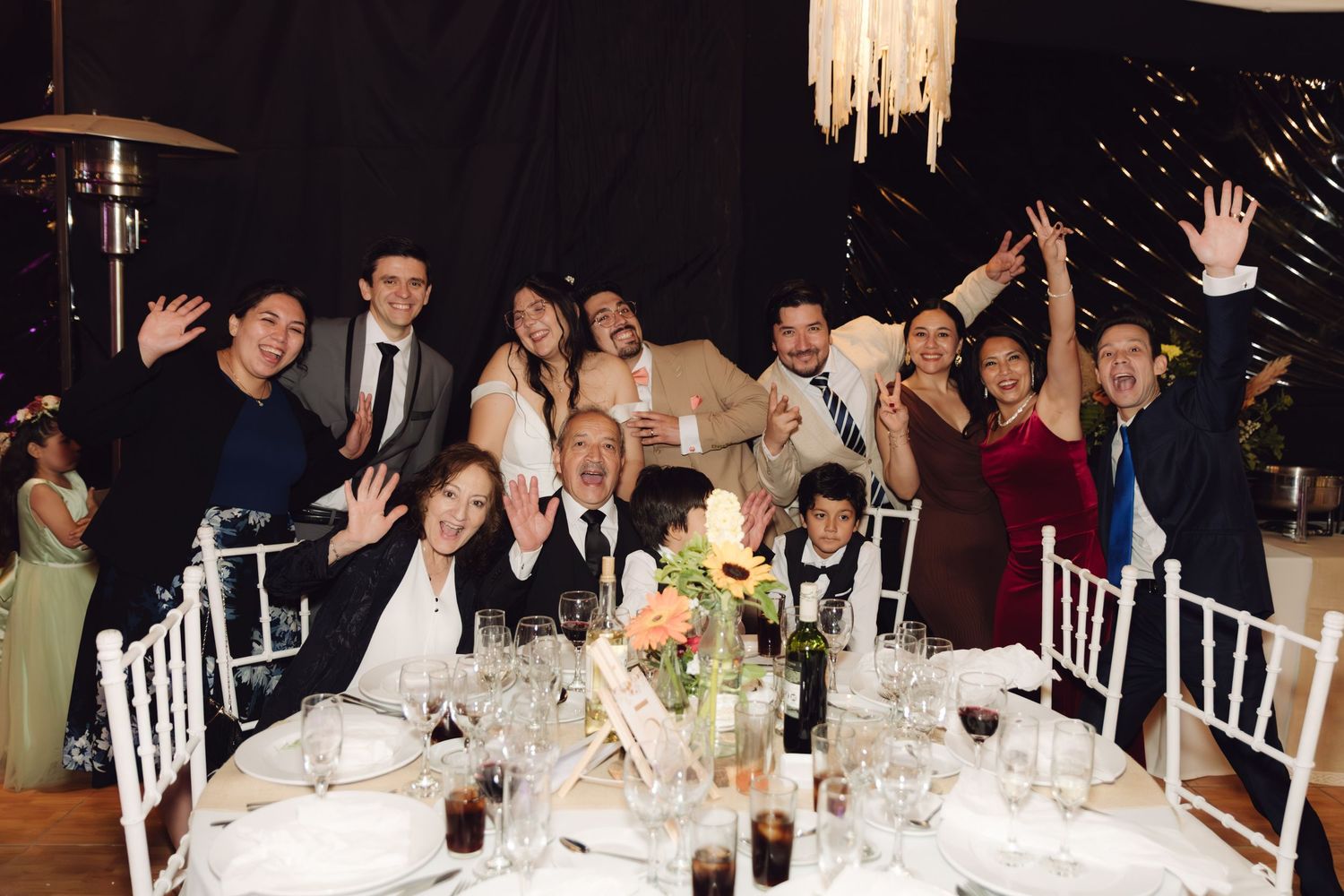 Cheerful wedding guests raising hands for a group photo at a formal reception dinner table.