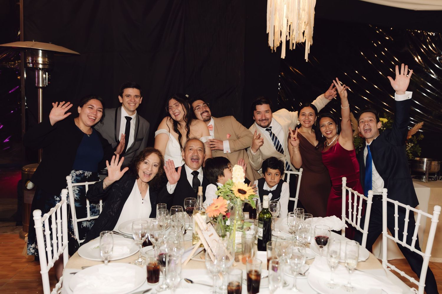 Smiling wedding guests waving at camera during a festive outdoor evening reception dinner.