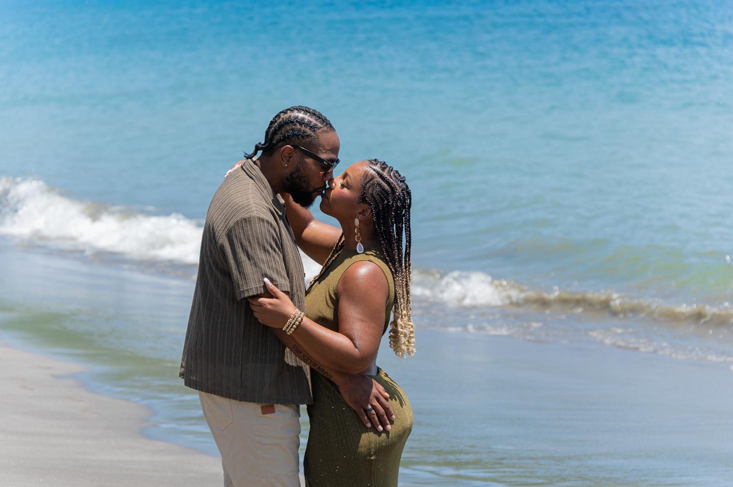 Couple sharing a romantic kiss on a sunny beach with turquoise ocean waves in the background.