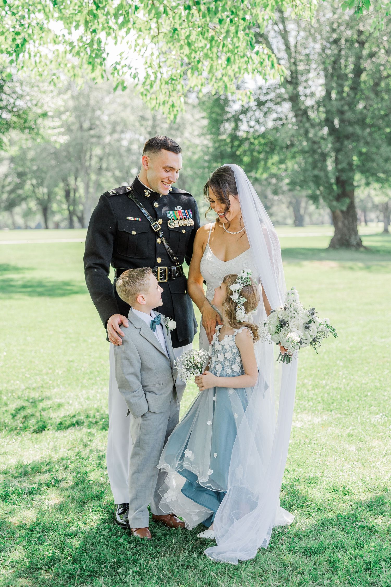 Military groom in dress uniform stands with bride in white gown and two children in a sunny green park setting.