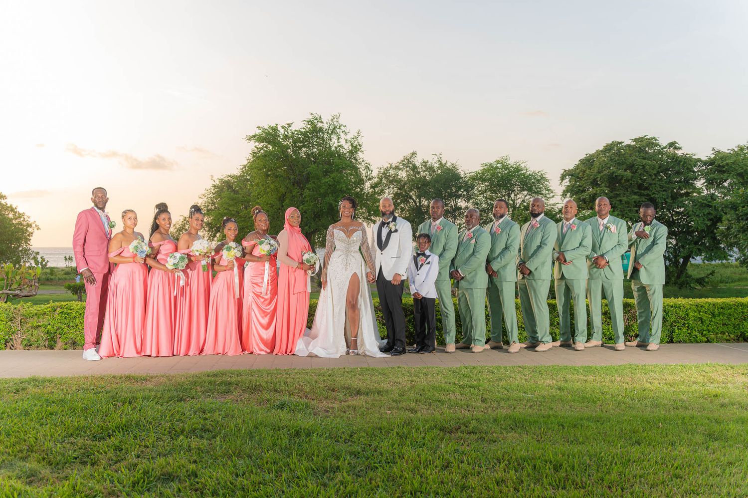 Wedding party poses outdoors at sunset, bridesmaids in coral gowns and groomsmen in sage green suits flanking the bride and groom.