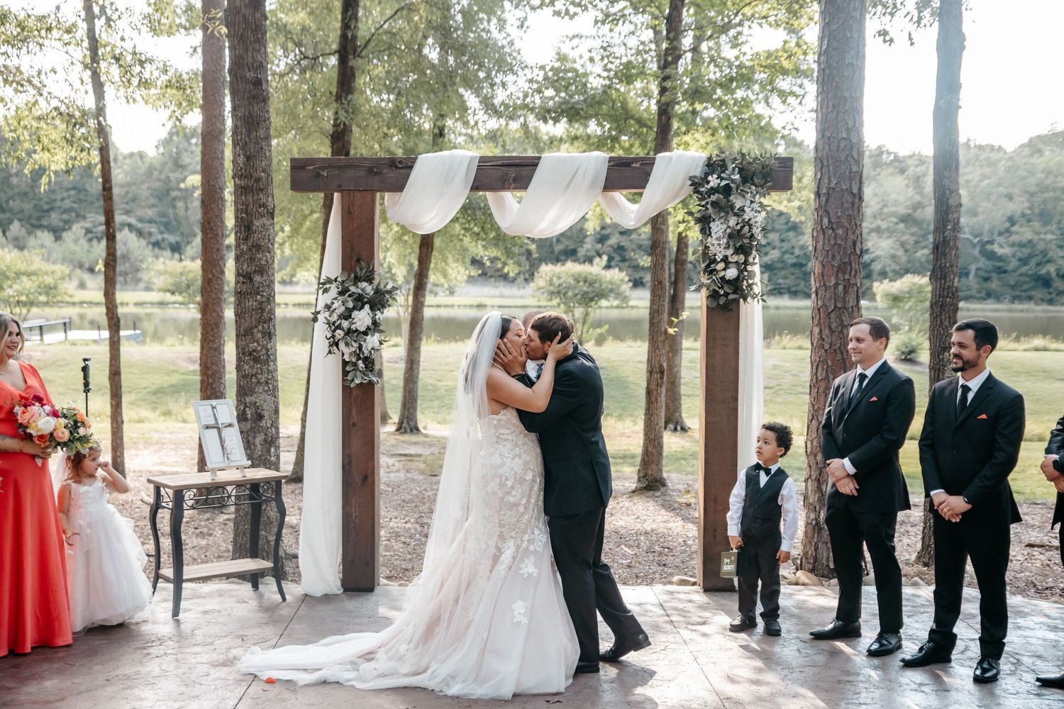 Bride and groom share first kiss at outdoor wedding ceremony under rustic wooden arch with white draping and floral decor.