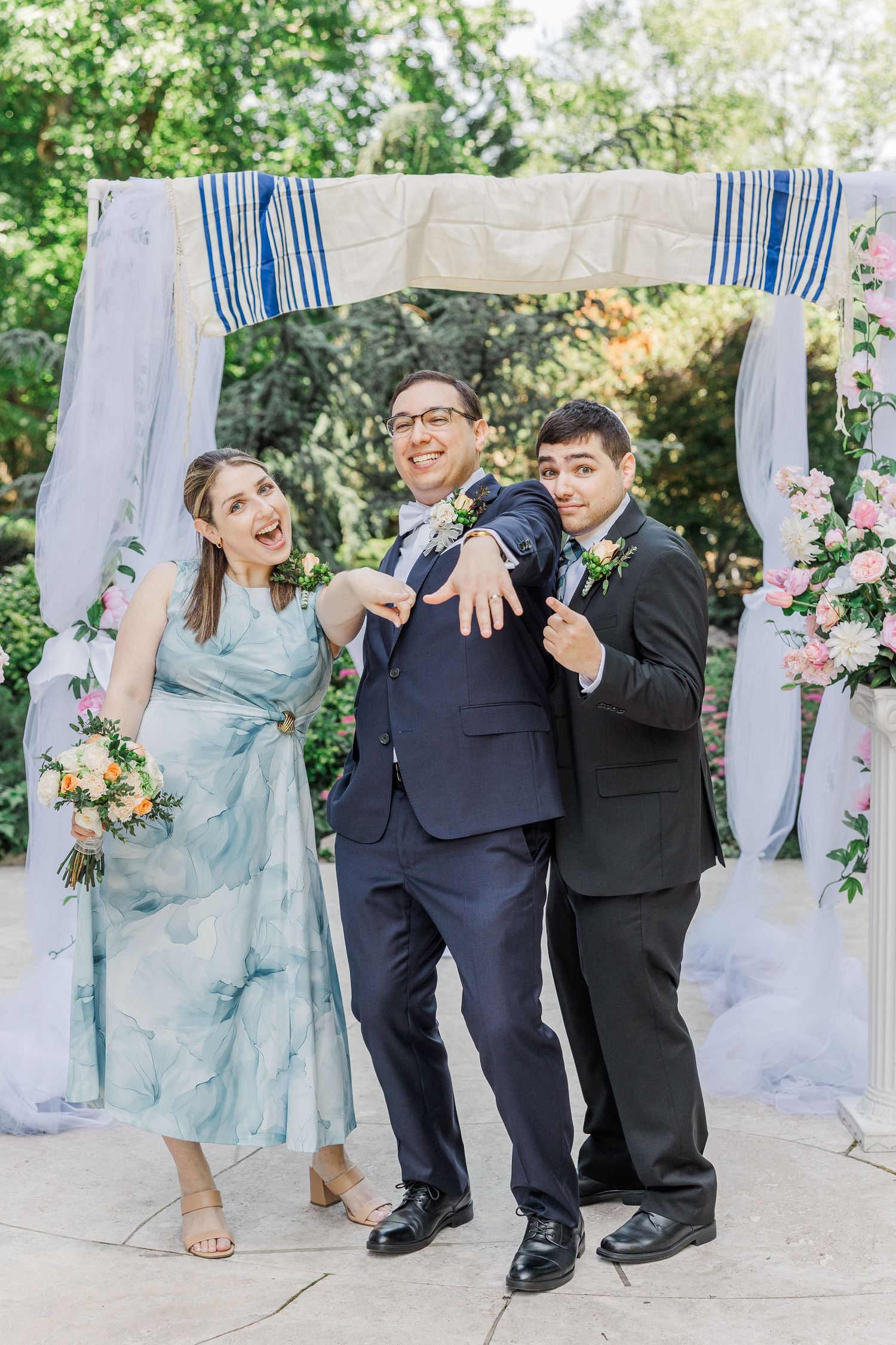 Three people pose joyfully under a floral chuppah at a Jewish wedding ceremony outdoors.