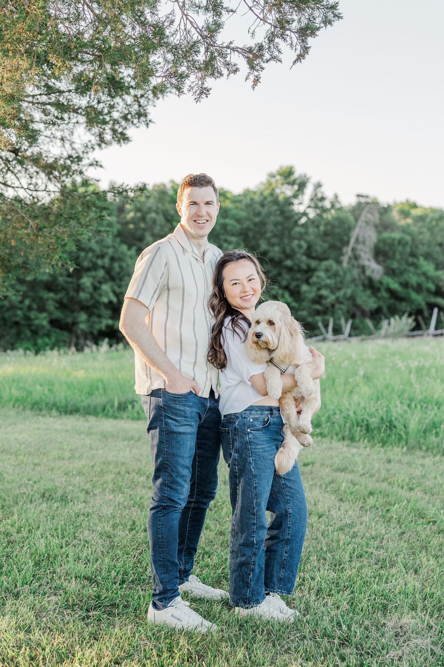 A smiling couple stands outdoors on green grass, the woman holding a fluffy dog, with trees in the background.