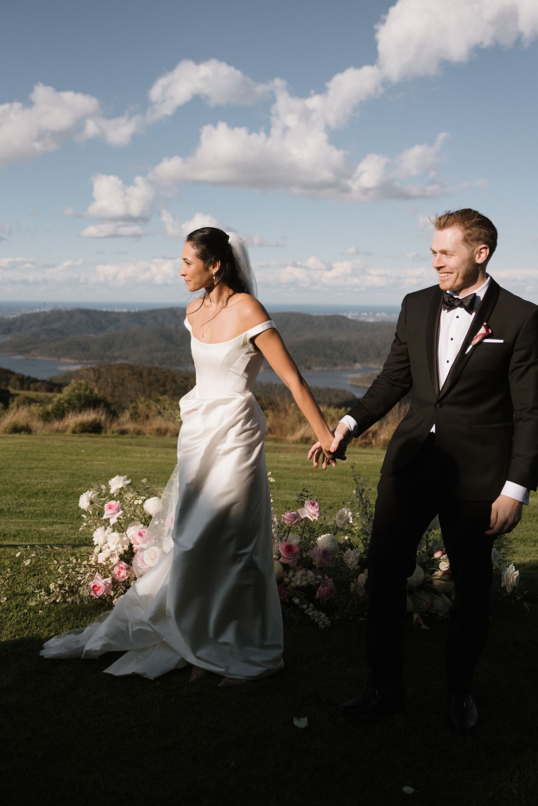 Bride in off-shoulder gown and groom in tuxedo hold hands at outdoor wedding with scenic hills backdrop.