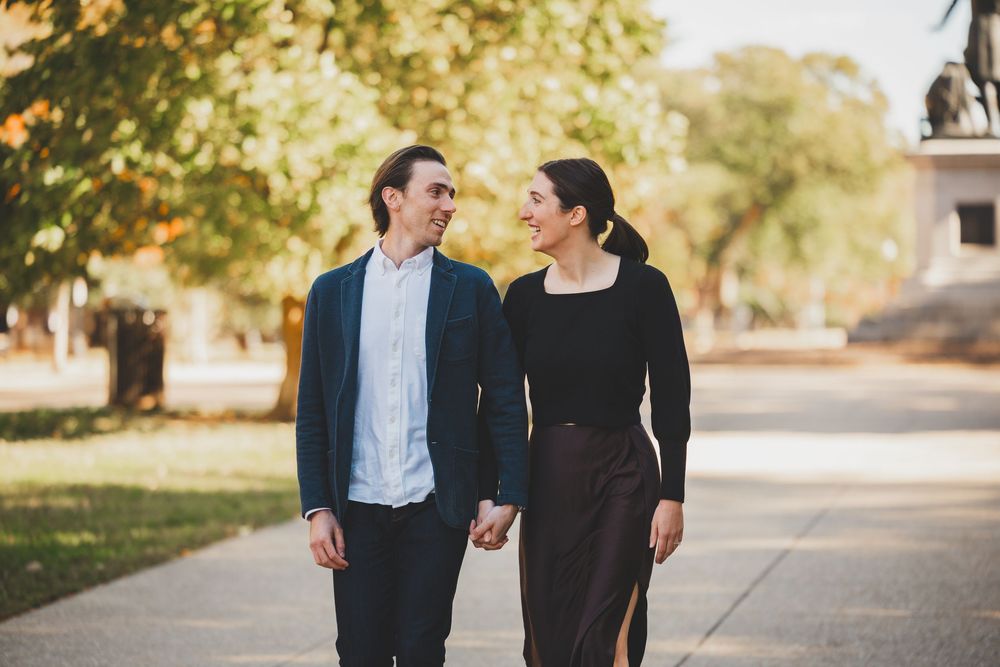 Couple holding hands and smiling at each other while walking through a tree-lined park path.