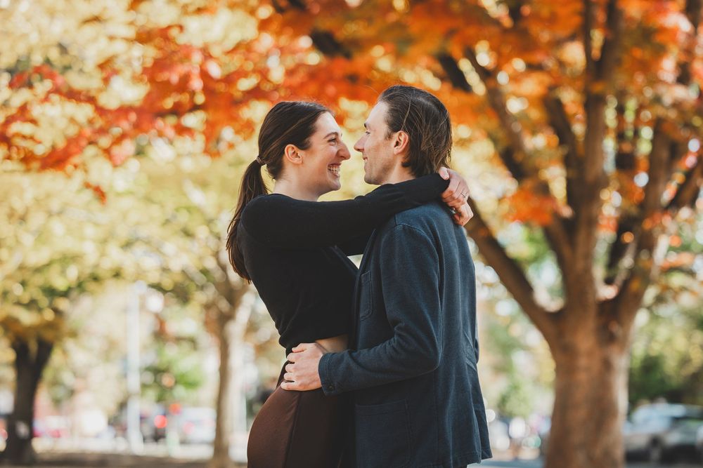 Couple embracing and smiling at each other outdoors among vibrant autumn orange and red trees at Lincoln Park in Washington DC