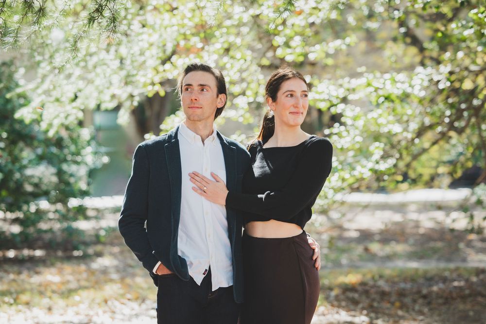 A couple poses outdoors among blooming spring trees, man in navy suit, woman in black top and skirt.