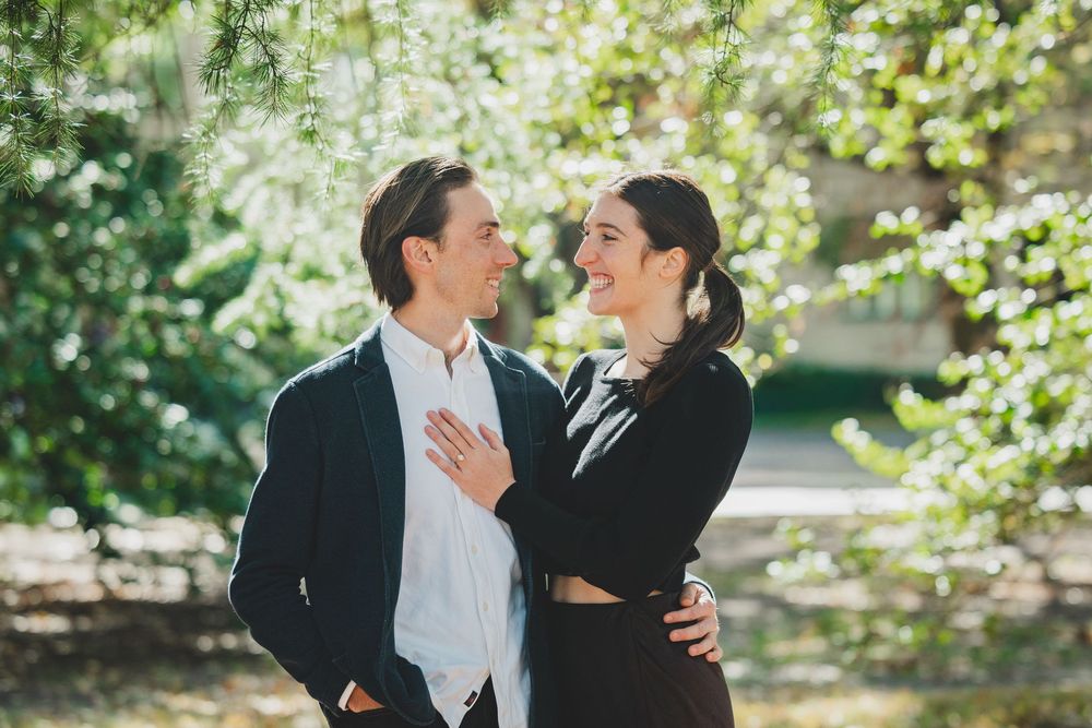 A smiling couple in elegant black attire share a romantic moment outdoors among lush green trees.