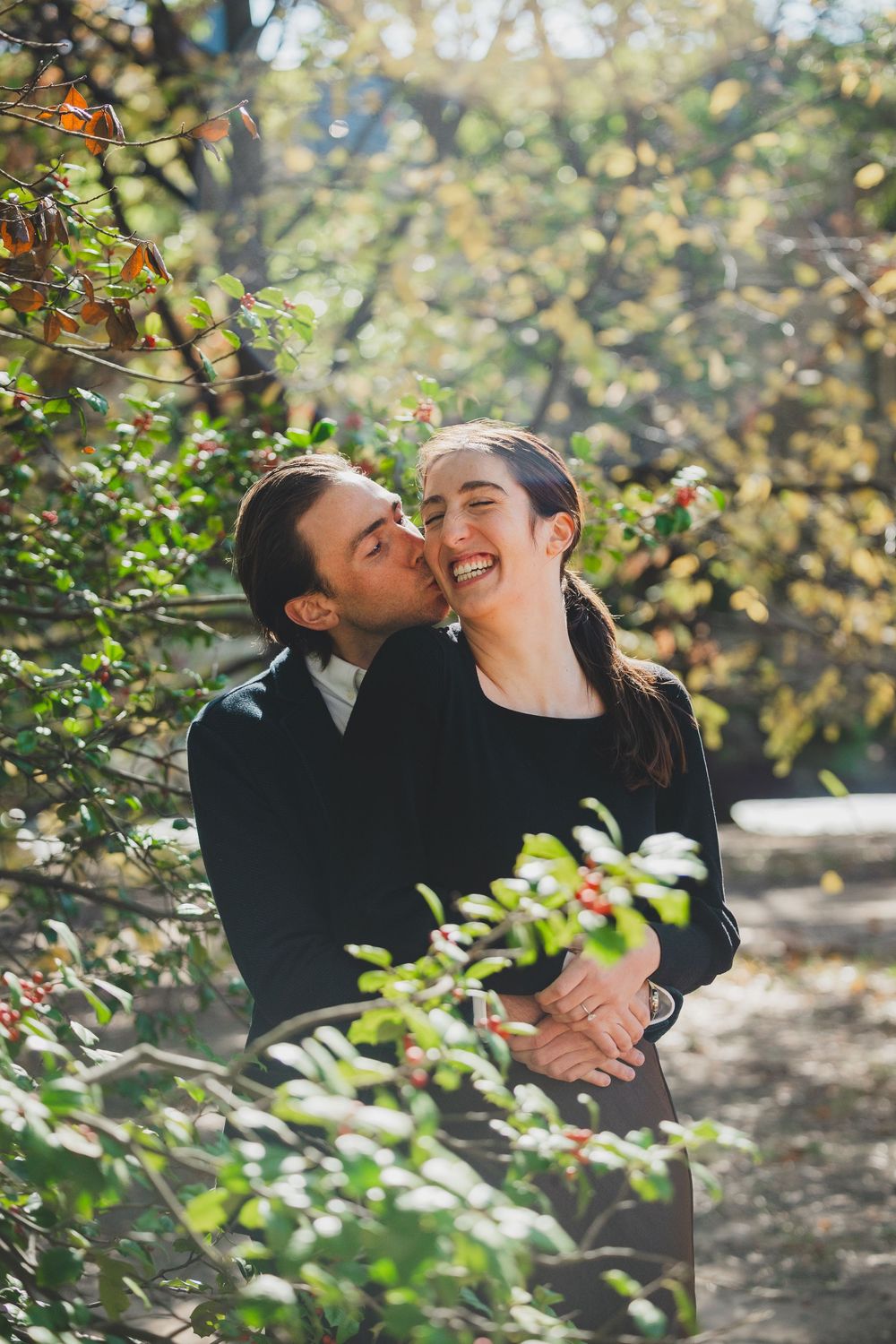 A couple shares a tender kiss outdoors, surrounded by lush green foliage in warm sunlight.