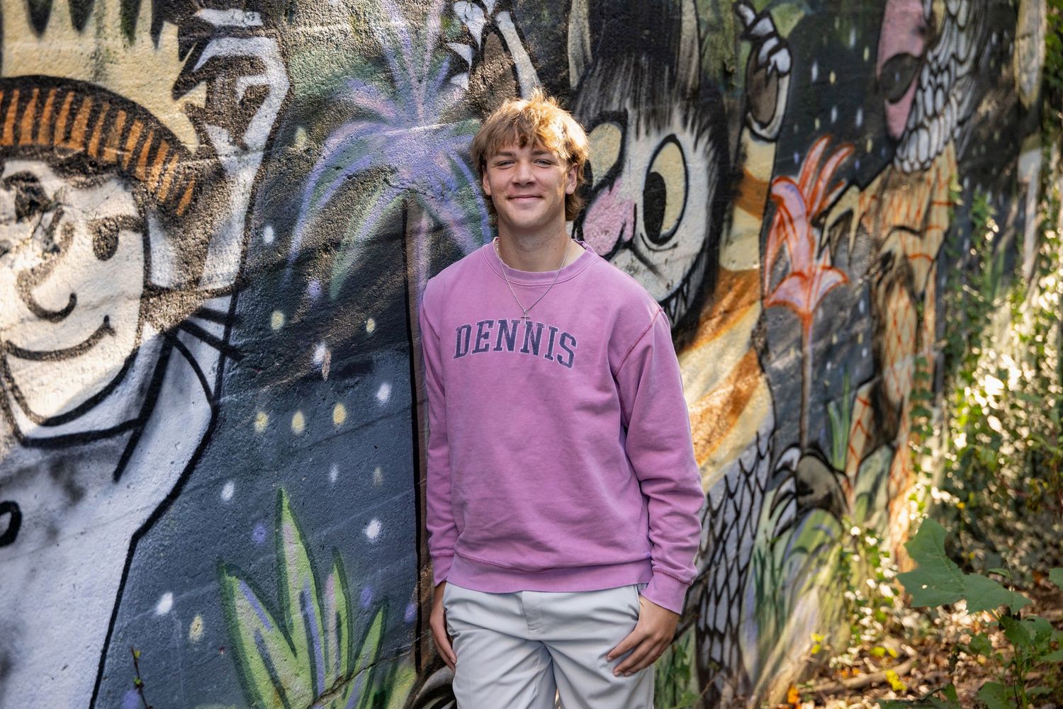 Young man in pink Dennis sweatshirt smiling in front of colorful graffiti mural wall outdoors.