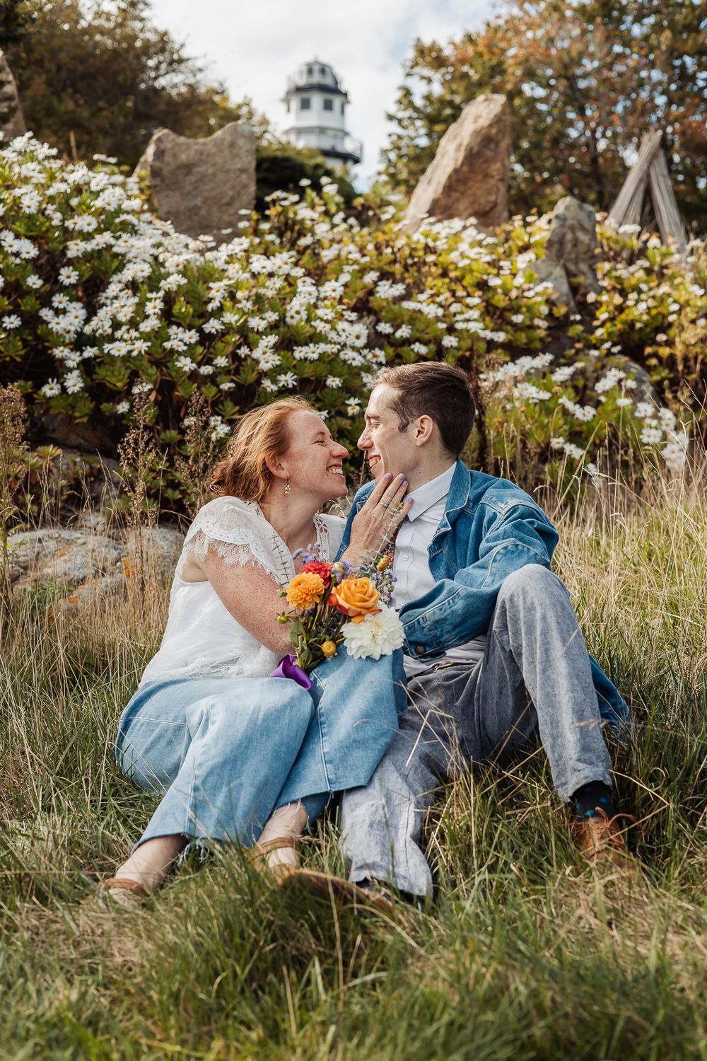 Couple sitting in grass near white wildflowers and lighthouse, sharing romantic moment with colorful bouquet.