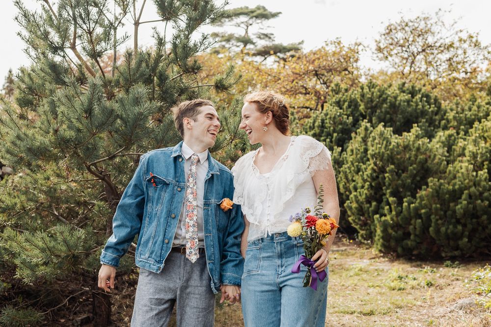 Couple holding hands and smiling at each other outdoors, woman holding colorful flowers, surrounded by green trees.