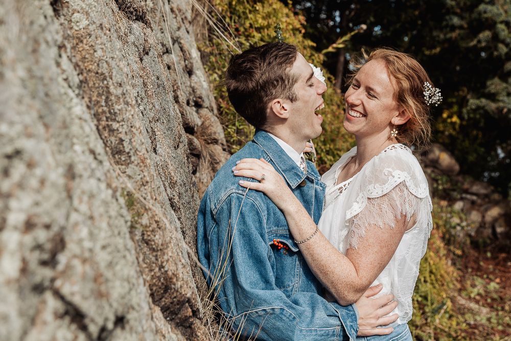 Couple embracing and smiling near a rocky wall outdoors, wearing casual attire in a natural setting.
