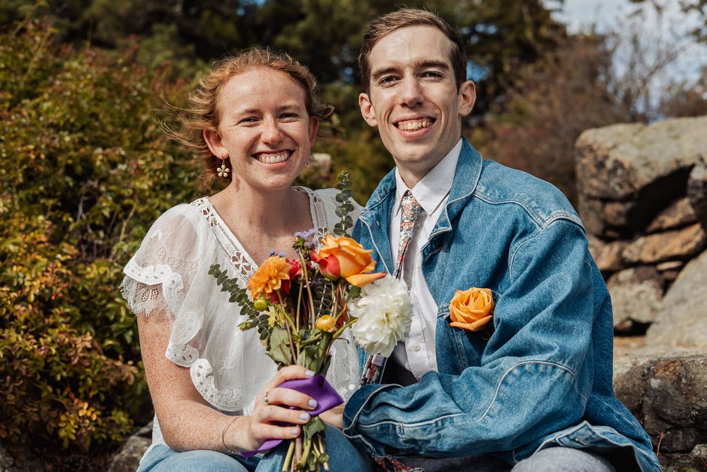 Smiling couple outdoors holding orange and white flower bouquet, woman in white dress, man in denim jacket with rose boutonniere.