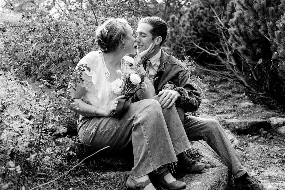Black and white photo of a romantic couple kissing while seated on rocks surrounded by lush greenery outdoors.