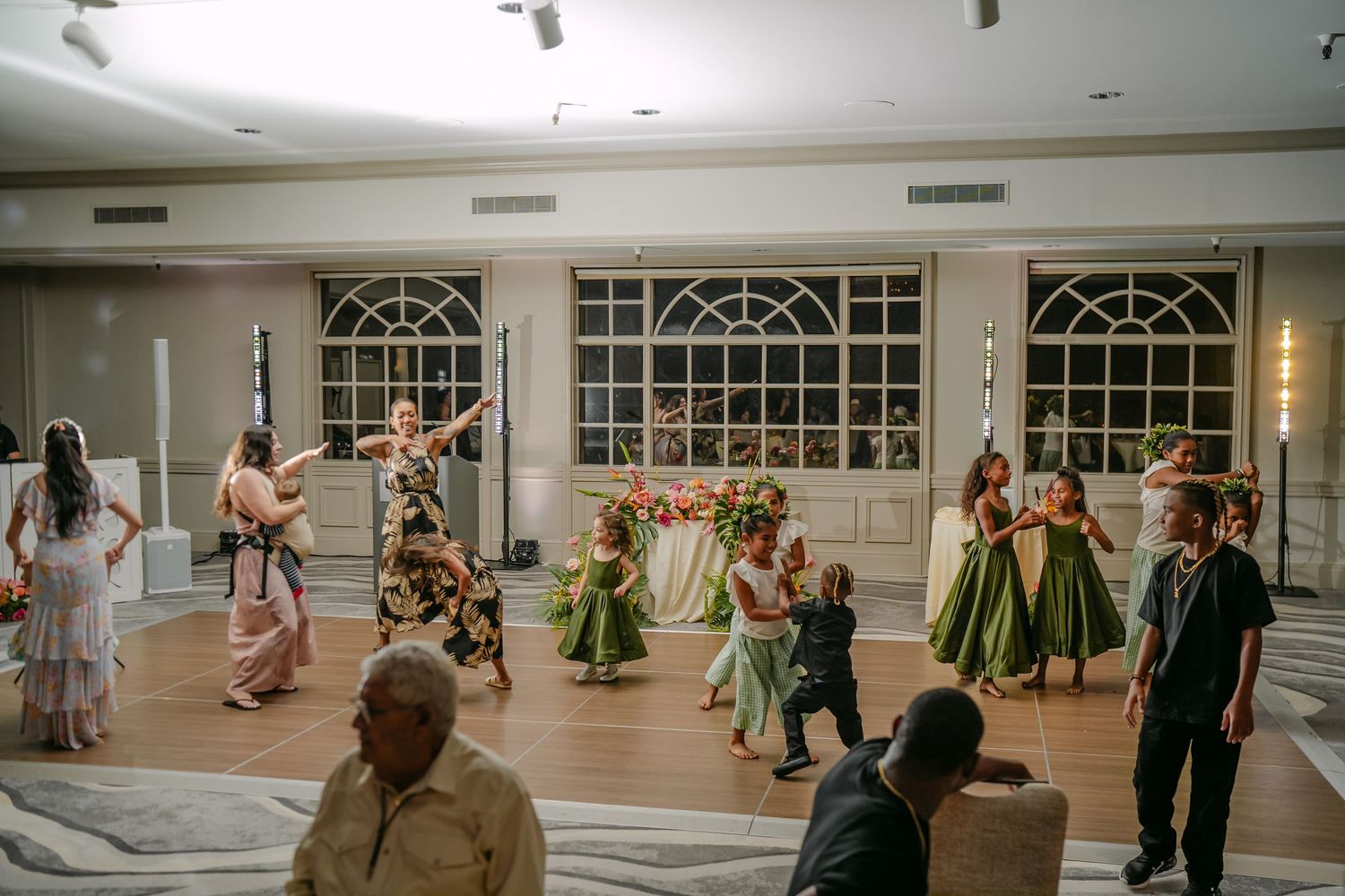 Hula dancers in green dresses perform at an elegant indoor wedding reception with large windows.